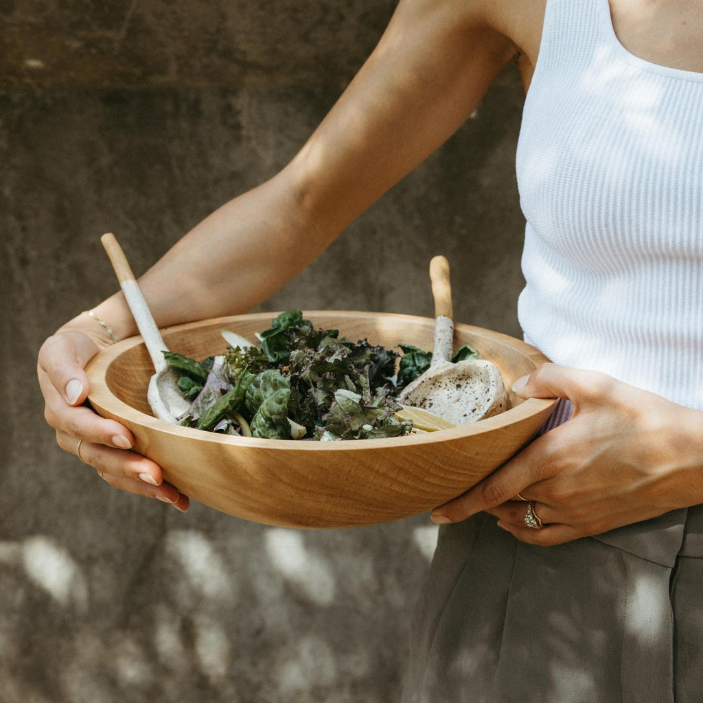 woman holding large wood salad bowl with salad and ceramic serving spoons