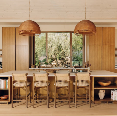 A modern kitchen interior with a central island, four woven bar stools, two large woven pendant lights, wood cabinetry, and a window overlooking greenery outdoors.