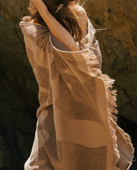 A person wrapped in a large brown beach towel, photographed outdoors against a dark rocky background.