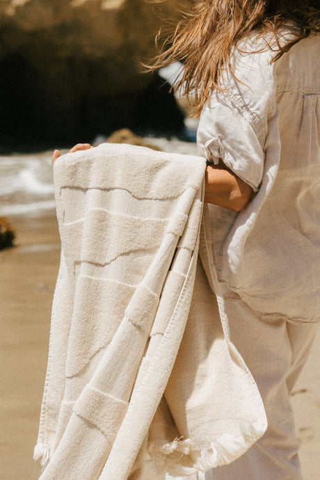 A person standing on a sandy beach holding a folded cream-colored cotton towel with stitched geometric patterns and fringed edges.